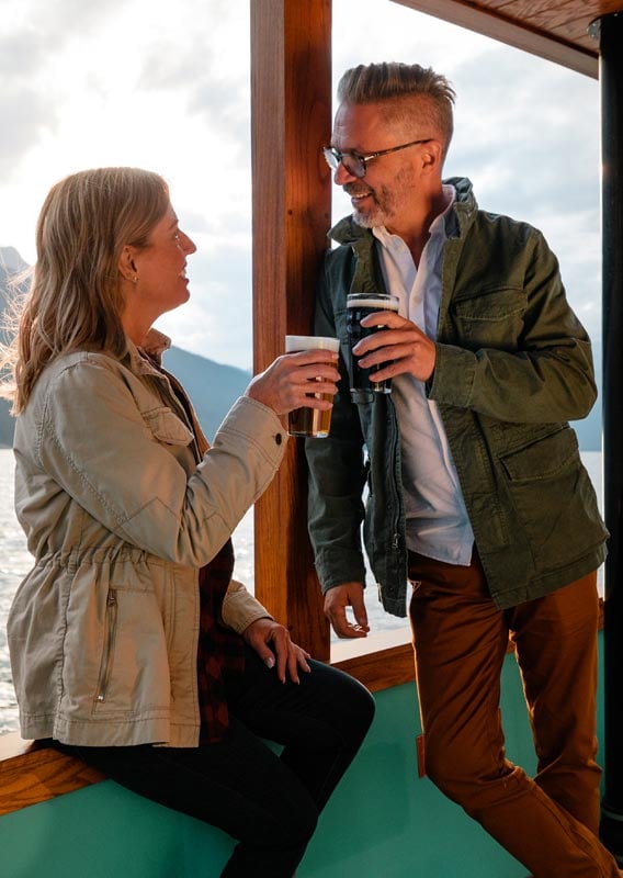 Two people hold glasses of beer in a cheers on the deck of a boat.