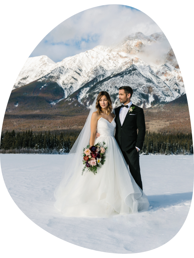Two brides hold hands at a rocky viewpoint above a small mountain lake.